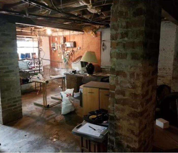 basement with water on the floor and a table with various items on it. Various furniture around and a window towards the back