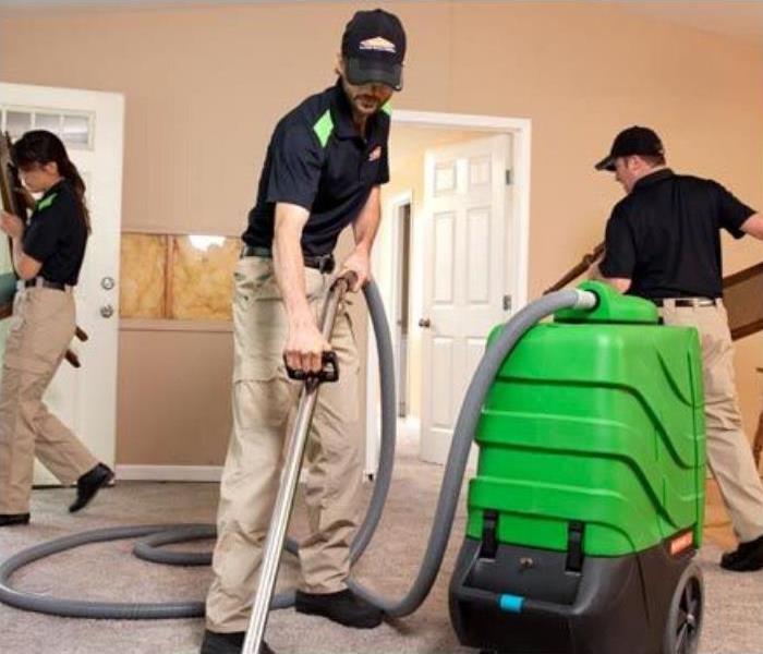 three people  wearing black polos and khaki cargo pants cleaning carpet with vacuum in house.